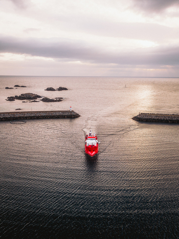 Aerial view of a vibrant red ship slicing through the tranquil dark waters toward the horizon framed by stone breakwaters Ballstad Nordland Norway. Aerial view of a vibrant red ship slicing through the tranquil dark waters toward the horizon framed by stone breakwaters Ballstad Nordland Norway.