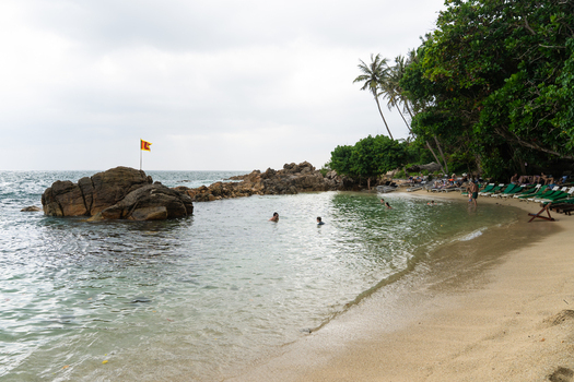 View of Secret Beach Mirissa with people swimming in clear water rocky formations with a flag and sun loungers under palm trees in Mirissa Southern Province Sri Lanka. View of Secret Beach Mirissa with people swimming in clear water rocky formations with a flag and sun loungers under palm trees in Mirissa Southern Province Sri Lanka.