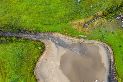 Aerial view of a muddy pond surrounded by lush green fields with hay bales and a small boat in Latvia.