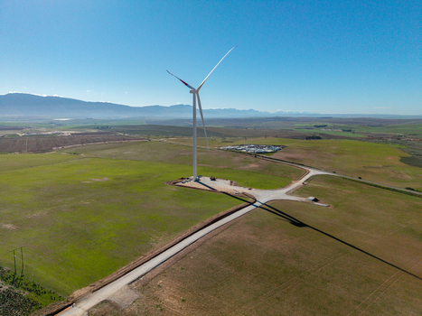 Aerial view of a large white wind turbine standing in a vast rural landscape with green fields and distant mountains under a clear blue sky in Swellendam Western Cape South Africa. Aerial view of a large white wind turbine standing in a vast rural landscape with green fields and distant mountains under a clear blue sky in Swellendam Western Cape South Africa.