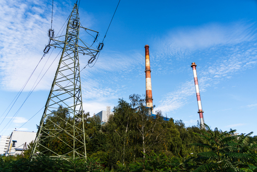 View of a power plant with tall chimneys and an electricity pylon surrounded by trees under a blue sky in Poland.