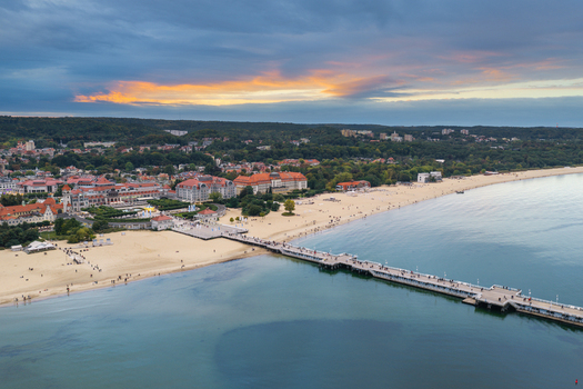 Aerial view of Sopot Pier extending into the Baltic Sea with sandy beach historic buildings and lush greenery under a dramatic sunset sky in Sopot Pomeranian Voivodeship Poland. Aerial view of Sopot Pier extending into the Baltic Sea with sandy beach historic buildings and lush greenery under a dramatic sunset sky in Sopot Pomeranian Voivodeship Poland.
