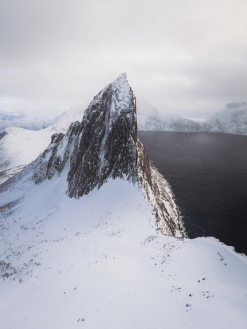 Aerial view of stark snow laden peaks rising majestically against the dark tranquil waters of the fjord a contrast of textures and tones under a somber sky Fjordgård Troms Norway. Aerial view of stark snow laden peaks rising majestically against the dark tranquil waters of the fjord a contrast of textures and tones under a somber sky Fjordgård Troms Norway.