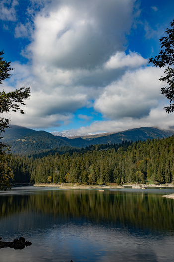 View of the Swiss Alps reflected in a calm lake surrounded by dense pine forests under a cloudy blue sky in Airolo Ticino Switzerland. View of the Swiss Alps reflected in a calm lake surrounded by dense pine forests under a cloudy blue sky in Airolo Ticino Switzerland.