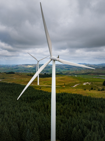 Aerial view of Hog Jaeren windkraftverk featuring large white wind turbines standing above a dense coniferous forest and rolling green hills in Ha Municipality Rogaland Norway.
