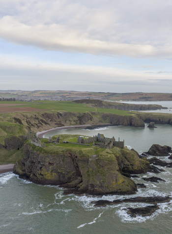 Aerial view of the medieval ruins of Dunnottar Castle perched on a dramatic cliff edge overlooking the North Sea under a cloudy sky in Stonehaven Scotland United Kingdom. Aerial view of the medieval ruins of Dunnottar Castle perched on a dramatic cliff edge overlooking the North Sea under a cloudy sky in Stonehaven Scotland United Kingdom.
