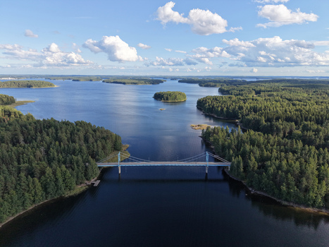 Aerial view of Potkusalmen silta Bridge crossing the calm waters of a lake surrounded by dense pine forests under a bright blue sky in Savonlinna Etela Savo Finland.