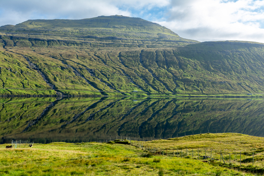 View of a mountain reflected in the calm waters of a fjord with lush green grassy slopes and a fence in the foreground in the Faroe Islands. View of a mountain reflected in the calm waters of a fjord with lush green grassy slopes and a fence in the foreground in the Faroe Islands.