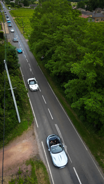 Aerial view of a convoy of luxury sports cars with people driving along a narrow road through lush green trees and vineyards in La Chapelle de Guinchay Bourgogne Franche Comte France.