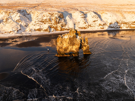 Aerial view of the Hvitserkur rock formation rising from the dark waters of the coast surrounded by snow covered cliffs under the warm light of the golden hour in Iceland. Aerial view of the Hvitserkur rock formation rising from the dark waters of the coast surrounded by snow covered cliffs under the warm light of the golden hour in Iceland.