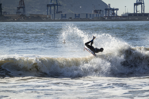 View of a bodyboarder performing an aerial maneuver on a breaking wave at Ereaga Beach with industrial port cranes in the background Algorta Euskadi Spain.