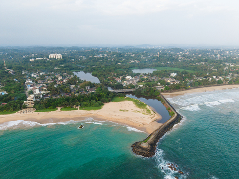 Aerial view of a stone breakwater extending into the turquoise sea next to a sandy beach and lush tropical greenery in Galle Southern Province Sri Lanka.