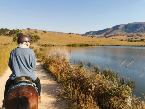 View of a person horseback riding on a dirt path beside a calm lake with grassy hills and mountains in the background in eManzana Mpumalanga South Africa. View of a person horseback riding on a dirt path beside a calm lake with grassy hills and mountains in the background in eManzana Mpumalanga South Africa.