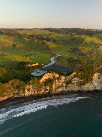 Aerial view of the rugged coastline where the river meets the sea cliffs bathed in golden light contrasting with the deep blue ocean Waikawau Waikato Region New Zealand. Aerial view of the rugged coastline where the river meets the sea cliffs bathed in golden light contrasting with the deep blue ocean Waikawau Waikato Region New Zealand.