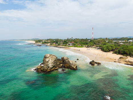 Aerial view of a large rock formation in turquoise water near a sandy beach with palm trees and a communication tower in Koggala Southern Province Sri Lanka. Aerial view of a large rock formation in turquoise water near a sandy beach with palm trees and a communication tower in Koggala Southern Province Sri Lanka.