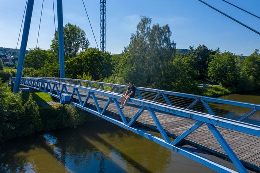Aerial view of the blue cable stayed bridge at Sportinsel Forchheim with a man sitting on the railing surrounded by lush green trees and calm water Forchheim Bayern Germany.