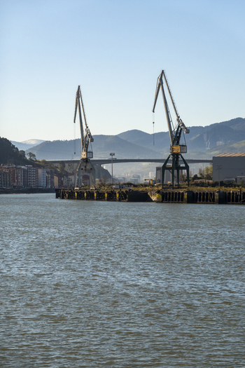 View of industrial cranes standing on the waterfront of the Nervion river with mountains and a bridge in the background in Greater Bilbao Basque Country Spain. View of industrial cranes standing on the waterfront of the Nervion river with mountains and a bridge in the background in Greater Bilbao Basque Country Spain.