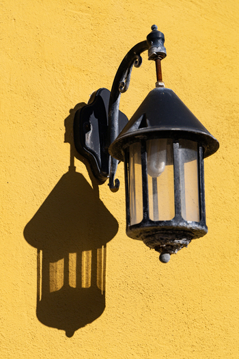 View of a vintage black outdoor lantern casting a sharp shadow on a bright yellow textured wall in Ribe Denmark. View of a vintage black outdoor lantern casting a sharp shadow on a bright yellow textured wall in Ribe Denmark.