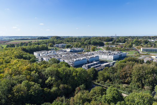 Aerial view of a large data center complex surrounded by dense green forest under a clear blue sky in United Kingdom.