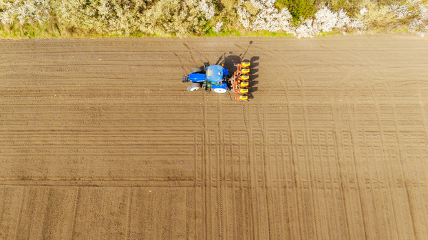 Aerial view of a blue tractor sowing seeds in a brown field with a line of white blossoming trees in the background Sremska Mitrovica Vojvodina Serbia. Aerial view of a blue tractor sowing seeds in a brown field with a line of white blossoming trees in the background Sremska Mitrovica Vojvodina Serbia.