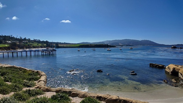 View of 17 Mile Drive featuring a wooden pier extending into the calm bay with sailboats rocky shoreline and green hills in Del Monte Forest California United States. View of 17 Mile Drive featuring a wooden pier extending into the calm bay with sailboats rocky shoreline and green hills in Del Monte Forest California United States.
