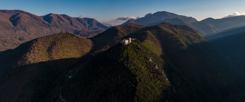 Aerial view of the Santuario del Santissimo Salvatore perched on a mountain peak surrounded by lush green hills and peaks under a clear sky in Montella Campania Italy. Aerial view of the Santuario del Santissimo Salvatore perched on a mountain peak surrounded by lush green hills and peaks under a clear sky in Montella Campania Italy.