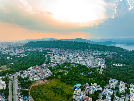 Aerial view of the urban landscape of Udaipur featuring dense housing lush green trees a prominent hill and a distant lake under a dramatic sunset sky in Udaipur Rajasthan India. Aerial view of the urban landscape of Udaipur featuring dense housing lush green trees a prominent hill and a distant lake under a dramatic sunset sky in Udaipur Rajasthan India.