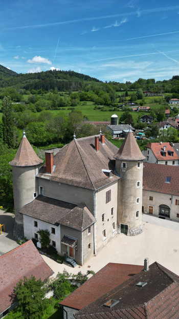 Aerial view of the historic Chateau De Vesancy featuring round stone towers and tiled roofs surrounded by lush green hills and a small village in Vesancy Auvergne Rhone Alpes France. Aerial view of the historic Chateau De Vesancy featuring round stone towers and tiled roofs surrounded by lush green hills and a small village in Vesancy Auvergne Rhone Alpes France.