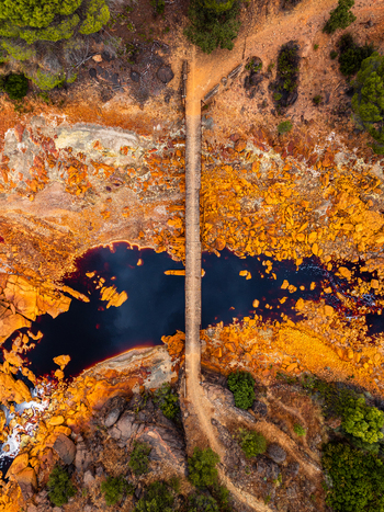 Aerial view of a narrow bridge crossing a dark mineral rich river surrounded by vibrant orange and yellow rocky terrain and sparse vegetation in Andalusia Spain. Aerial view of a narrow bridge crossing a dark mineral rich river surrounded by vibrant orange and yellow rocky terrain and sparse vegetation in Andalusia Spain.