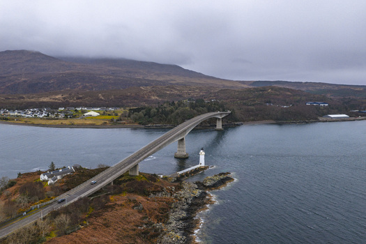 Aerial view of the Skye Bridge and Kyleakin Lighthouse spanning the sea with rolling hills and a cloudy sky in Isle of Skye Scotland United Kingdom.