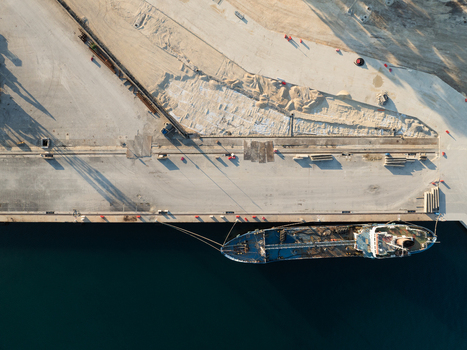 Aerial view of a large blue cargo ship moored at a concrete industrial pier with construction materials and long shadows in Augusta Sicily Italy. Aerial view of a large blue cargo ship moored at a concrete industrial pier with construction materials and long shadows in Augusta Sicily Italy.