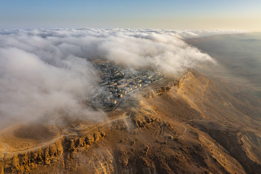 Aerial view of Mitzpe Ramon town situated on the edge of the Makhtesh Ramon crater with clouds rolling over the desert landscape during golden hour in Mitzpe Ramon South District Israel. Aerial view of Mitzpe Ramon town situated on the edge of the Makhtesh Ramon crater with clouds rolling over the desert landscape during golden hour in Mitzpe Ramon South District Israel.