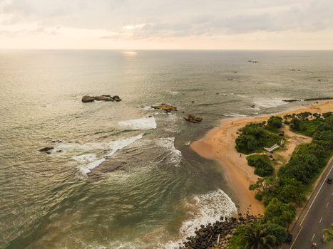 Aerial view of Dadalla beach with waves crashing on the sandy shore and a coastal road lined with trees under a soft sunset sky in Galle Southern Province Sri Lanka. Aerial view of Dadalla beach with waves crashing on the sandy shore and a coastal road lined with trees under a soft sunset sky in Galle Southern Province Sri Lanka.