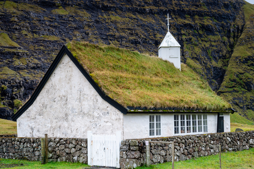 View of a traditional church with a grass roof white walls and a stone fence nestled against a steep dark grassy mountain slope in the Faroe Islands.