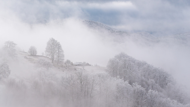 View of a snow covered mountain landscape with frost laden trees and a small house shrouded in thick morning fog in Alba Iulia Alba County Romania. View of a snow covered mountain landscape with frost laden trees and a small house shrouded in thick morning fog in Alba Iulia Alba County Romania.