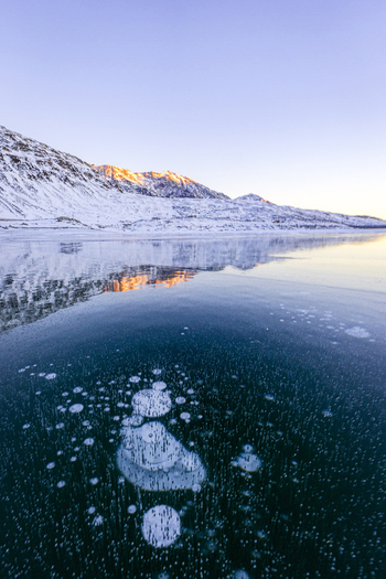 View of frozen lake with air bubbles trapped under the ice surface and snow covered mountains reflecting in the water at sunset in Madesimo Lombardy Italy. View of frozen lake with air bubbles trapped under the ice surface and snow covered mountains reflecting in the water at sunset in Madesimo Lombardy Italy.