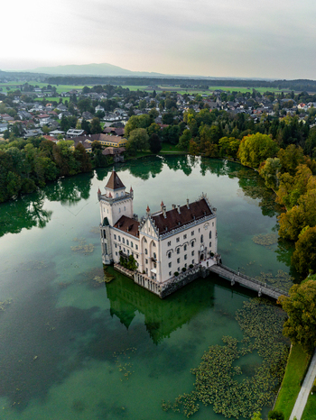 Aerial view of Schloss Anif surrounded by a calm lake with lush green trees and a wooden bridge in Anif Salzburg Austria.
