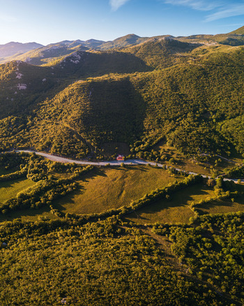 Aerial view of lush green mountains and a winding road passing by a small house under a clear blue sky in Rijeka Primorje Gorski Kotar Croatia. Aerial view of lush green mountains and a winding road passing by a small house under a clear blue sky in Rijeka Primorje Gorski Kotar Croatia.