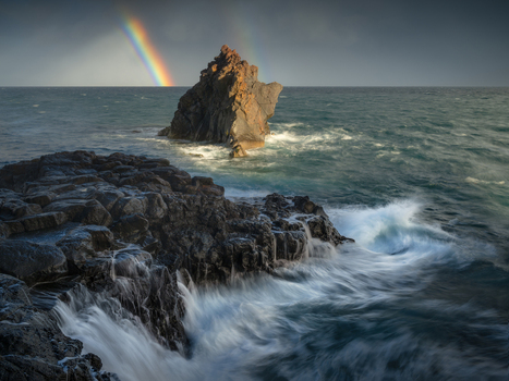 View of a dramatic volcanic rock formation rising from the Atlantic Ocean with waves crashing against dark basalt cliffs under a rainbow in Madeira Portugal. View of a dramatic volcanic rock formation rising from the Atlantic Ocean with waves crashing against dark basalt cliffs under a rainbow in Madeira Portugal.