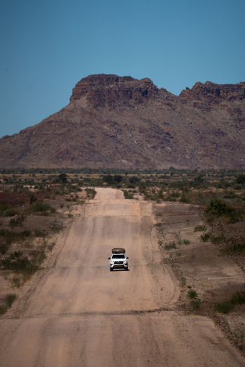 View of a lone white vehicle traversing a dusty sun baked road towards a massive rugged mountain under a clear blue sky Karas Region Namibia Keetmanshoop Karas Region Namibia. View of a lone white vehicle traversing a dusty sun baked road towards a massive rugged mountain under a clear blue sky Karas Region Namibia Keetmanshoop Karas Region Namibia.