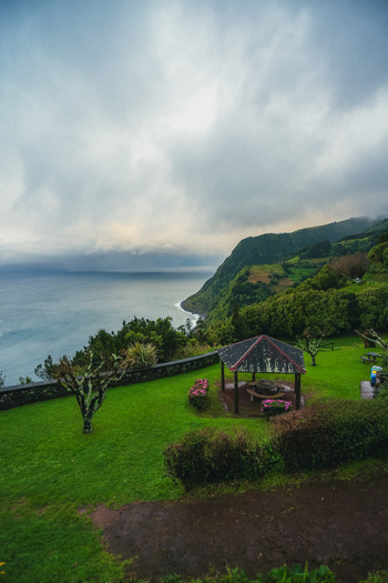 Aerial view of a rustic gazebo nestled on vibrant green grass overlooking the rugged coastline meeting the vast ocean under a cloudy sky Nordeste Azores Portugal.