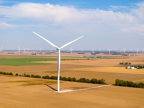 Aerial view of a large wind turbine in a vast agricultural field with numerous other turbines visible across the rural landscape under a clear blue sky in Wolcott Indiana United States.