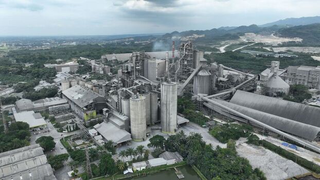 Aerial view of a large industrial factory complex with tall silos chimneys and conveyor belts surrounded by lush green trees and hills in Klapanunggal West Java Indonesia. Aerial view of a large industrial factory complex with tall silos chimneys and conveyor belts surrounded by lush green trees and hills in Klapanunggal West Java Indonesia.
