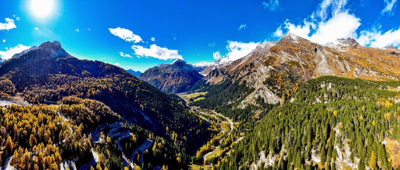 Aerial view of the winding Maloja Pass road surrounded by vibrant autumn forests and rugged mountain peaks under a clear blue sky in Bregaglia Grisons Switzerland. Aerial view of the winding Maloja Pass road surrounded by vibrant autumn forests and rugged mountain peaks under a clear blue sky in Bregaglia Grisons Switzerland.