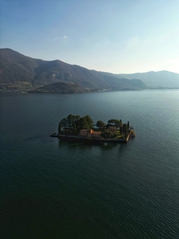 Aerial view of Isola di Loreto with its neo Gothic villa and lush gardens on Lake Iseo surrounded by calm water and distant mountains Monte Isola Lombardy Italy. Aerial view of Isola di Loreto with its neo Gothic villa and lush gardens on Lake Iseo surrounded by calm water and distant mountains Monte Isola Lombardy Italy.