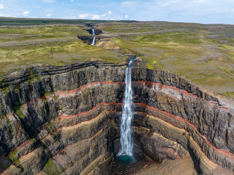 Aerial view of Hengifoss waterfall cascading over dramatic red and black basalt rock layers in the Icelandic highlands surrounded by green tundra under a clear blue sky Iceland. Aerial view of Hengifoss waterfall cascading over dramatic red and black basalt rock layers in the Icelandic highlands surrounded by green tundra under a clear blue sky Iceland.