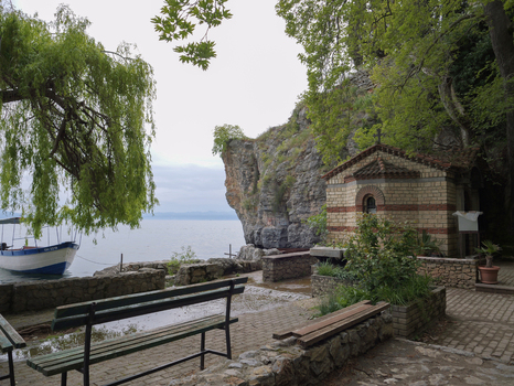View of the small stone chapel near rocky cliffs and Lake Ohrid with a wooden boat moored at the shore and a park bench in the foreground Ohrid Municipality of Ohrid North Macedonia. View of the small stone chapel near rocky cliffs and Lake Ohrid with a wooden boat moored at the shore and a park bench in the foreground Ohrid Municipality of Ohrid North Macedonia.