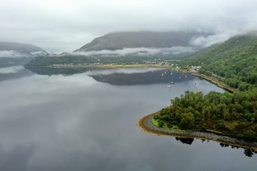 Aerial view of the calm waters of Loch Leven surrounded by mist covered mountains lush green forests and small boats moored near the shore in Glencoe Scotland United Kingdom.