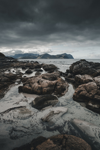 View of the rugged rocky shoreline of Vikten Beach with dark dramatic clouds over the distant mountains in Vikten Nordland Norway.