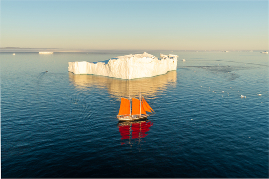 Aerial view of a sailboat with vibrant orange sails navigating calm blue waters near a massive white iceberg under a clear sky in Greenland. Aerial view of a sailboat with vibrant orange sails navigating calm blue waters near a massive white iceberg under a clear sky in Greenland.